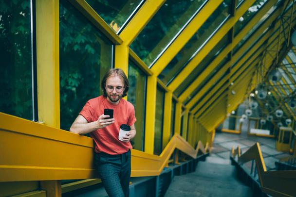 Young man concentrated on phone screen while standing against glass wall and leaning on railing and holding cup of coffee at enclosed pedestrian bridge - Photo, Image