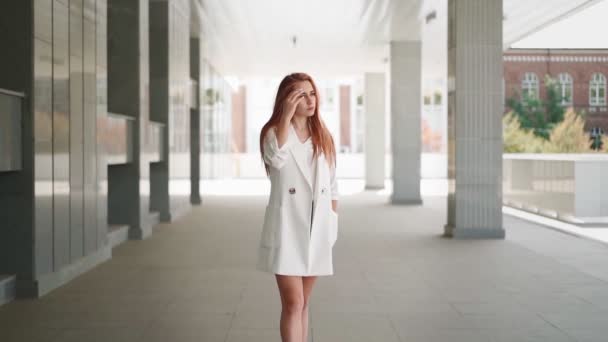Όμορφη σύγχρονη επιχειρηματική γυναίκα με μακριά κόκκινα μαλλιά σε μοντέρνο λευκό παλτό στέκεται κοντά σε ένα κτίριο γραφείων. Γυναίκα επιχειρηματίας με αυτοπεποίθηση. Ελκυστική επιτυχημένη κυρία με μοντέρνα ρούχα. Αργή κίνηση. - Πλάνα, βίντεο
