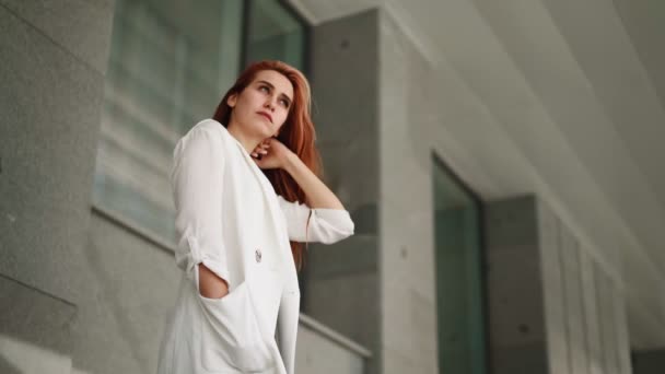 Beautiful modern business woman with long red hair in trendy white coat stands near an office building. Confident female entrepreneur. Attractive successful lady in fashionable clothes. Slow motion. - Footage, Video