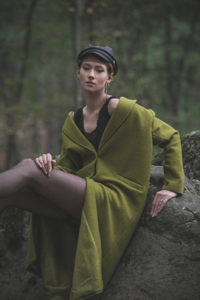 Fashionable young woman in green coat, cap and black tights posing in a forest on a stone. Art portrait - Фото, зображення