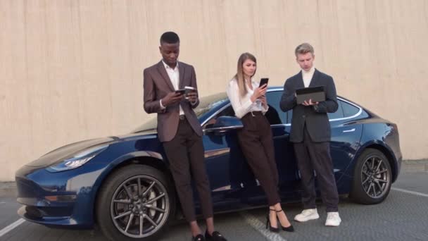 Drie jonge succesvolle mensen op de achtergrond van een sport elektrische auto, gebruik gadgets telefoon en tablet. technologie en mensen concept.Business mensen. Welvaart. - Video