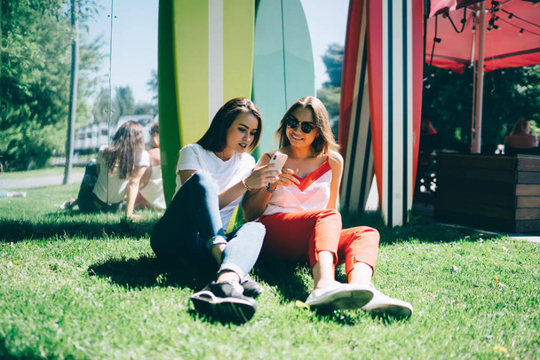 Millennial women resting at summer grass using cellular application for making together booking, youthful female bloggers checking web statistics of vlog popularity connected to 4g on smartphone - Фото, зображення