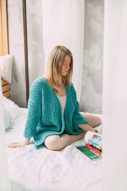 Młoda dziewczyna w piżamie i niebieskim swetrze siedzi na łóżku ze skrzyżowanymi nogami, przerzucając się przez magazyn. Dziewczyna cieszy się porankiem.. - Zdjęcie, obraz