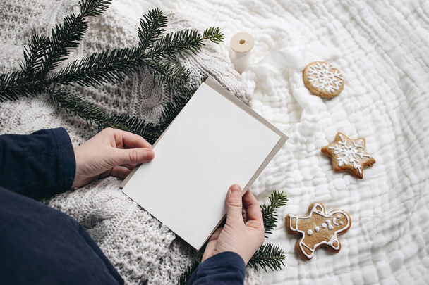 Christmas festive still life. Little child hands holding blank greeting card. Invitation mockup on knitted sweater. Gingerbread cookies and fir branches on white muslin blanket. Selective focus. - 写真・画像
