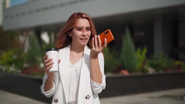Gyönyörű üzletasszony mosolyog és mobiltelefon hangfelvételt használ. Egy fiatal sikeres hölgy a vállalati iroda mellett, aki telefonon beszél. Modern női vállalkozó okostelefonnal. Lassú mozgás.. - Felvétel, videó