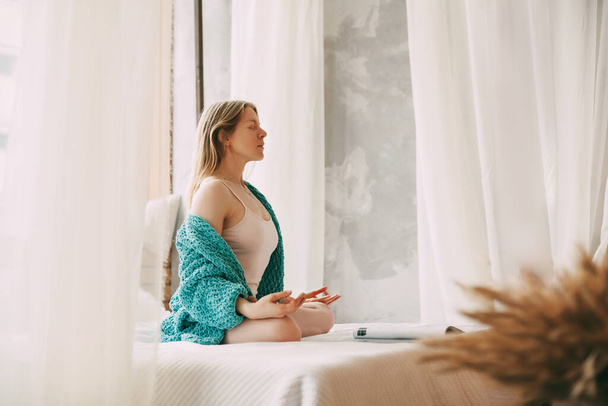 Μια νεαρή γυναίκα με πιτζάμες κάθεται σε ένα κρεβάτι σε μια θέση Lotus με τα δάχτυλά της διπλωμένα σε μια χειρονομία mudra, απολαμβάνοντας ένα πρωινό βαθύ διαλογισμό. Πλευρική άποψη. - Φωτογραφία, εικόνα