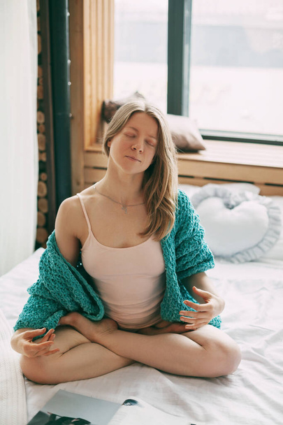 Une jeune fille médite en étant assise en position Lotus sur le lit dans la chambre. La fille fait du yoga anti-stress le matin à la maison. - Photo, image
