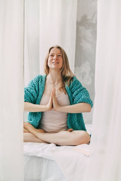 Uma jovem medita enquanto se senta na posição de Lótus na cama no quarto. A menina faz ioga anti-stress de manhã em casa. - Foto, Imagem