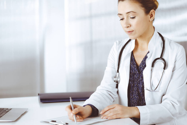 Cheerful intelligent woman-doctor is using a clipboard and writing some medication names, while sitting at the desk in her cabinet in a clinic. Portrait of professional female physician with a - 写真・画像