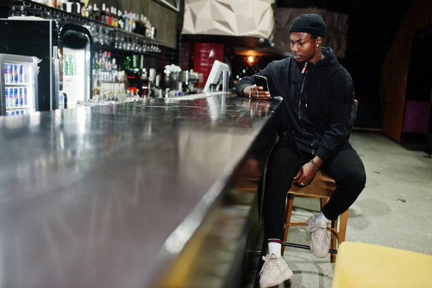 Handsome african american man posing  inside night club in black hat, sitting on bar counter. - Foto, afbeelding