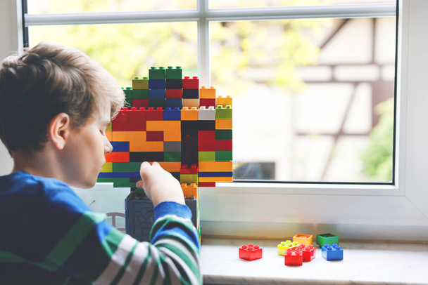 Little kid boy playing with lots of colorful plastic blocks. Adorable school child having fun with building and creating building by window. Creative leisure technic and robotic during corona time - Photo, Image