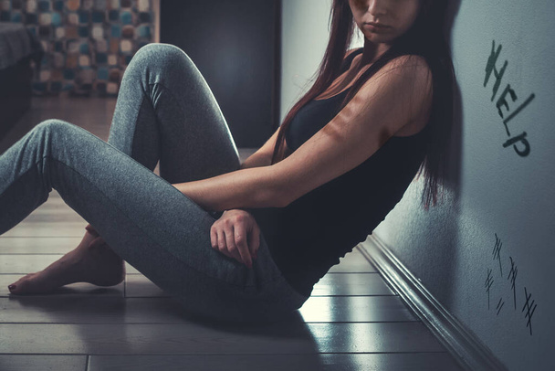Koncept domácího násilí. Smutná mladá žena s modřinou na podpaží a pod okem, sedící na podlaze v tmavé místnosti. V pozadí je okno se závěsem. Nápověda k nápisu . - Fotografie, Obrázek