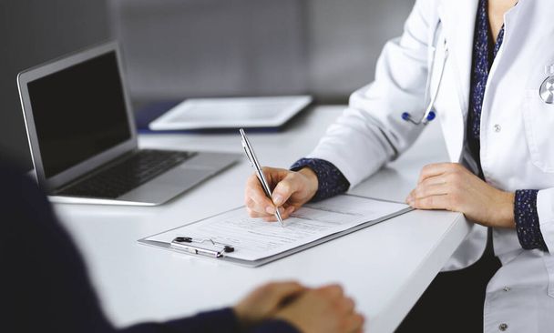 Unknown woman-doctor is writing some medical recommendations to her patient, while they are sitting together at the desk in the cabinet in a clinic. Physician is using a clipboard, close-up. Perfect - 写真・画像