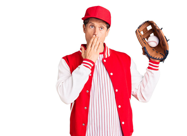 野球のユニフォームを着た若いハンサムな男がゴルフとボールを手で口をカバーし、ショックを受け、間違いを恐れている。驚きの表情  - 写真・画像