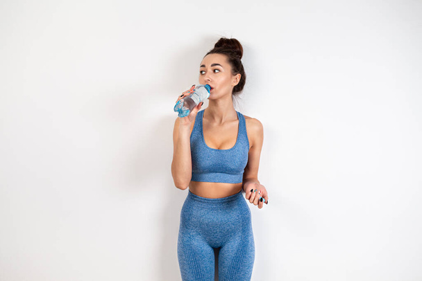 Het concept van gezond eten. Een jonge blanke vrouw in sportkleding, met een getinte figuur, die water drinkt uit een fles. Witte achtergrond. Kopieerruimte. - Foto, afbeelding