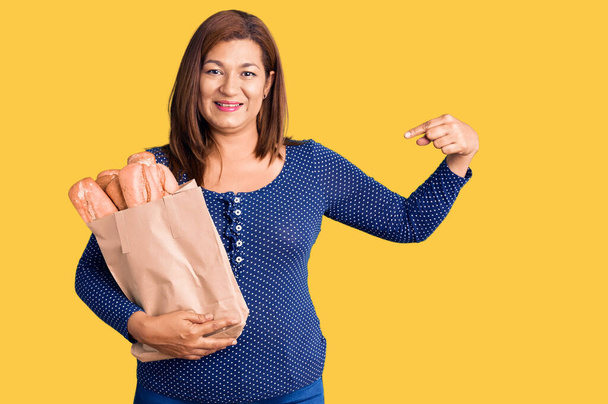 Латиноамериканка средних лет держит бумажный пакет с хлебом, указывая пальцем на себя, улыбаясь счастливой и гордой  - Фото, изображение