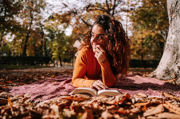 Μια όμορφη γυναίκα ξαπλωμένη σε μια κουβέρτα στο έδαφος με ένα βιβλίο σε μια υπέροχη ηλιόλουστη φθινοπωρινή ημέρα. Κοιτάζει κάτι περιέργως. Φθινοπωρινή έννοια του τρόπου ζωής στο ύπαιθρο - Φωτογραφία, εικόνα