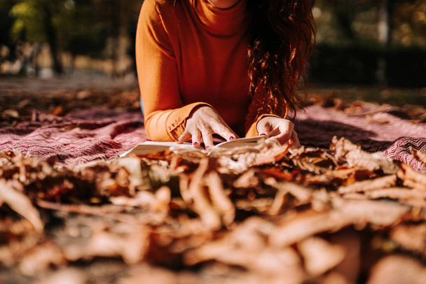 Ferme là. Concentration sélective. Une femme lisant un livre dans un parc un jour d'automne. Elle a des ongles marron et elle porte un pull orange. Mode de vie concept automnal - Photo, image