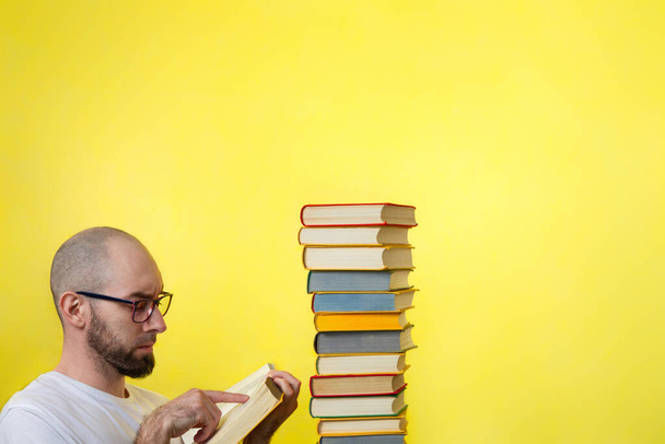 Concepto de educación. Un hombre calvo con barba y gafas, sosteniendo un libro y leyéndolo atentamente. Muy cerca hay una pila de libros. Fondo amarillo. Copiar espacio. - Foto, imagen