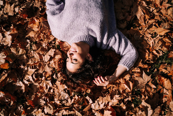 Una hermosa morena descansando tumbada en el suelo rodeada de hojas en un parque. Está disfrutando de un día soleado en otoño. Retrato otoñal y estilo de vida - Foto, Imagen