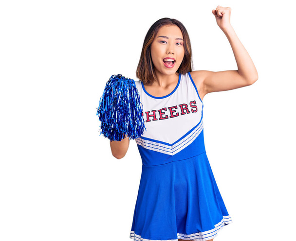 Young beautiful chinese girl wearing cheerleader uniform holding pompom screaming proud, celebrating victory and success very excited with raised arms  - Photo, Image