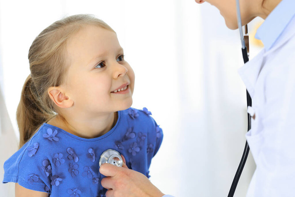 Az orvos sztetoszkóppal vizsgál meg egy kislányt. Boldog mosolygós gyerekbeteget a szokásos orvosi vizsgálaton. Orvosi és egészségügyi koncepciók - Fotó, kép