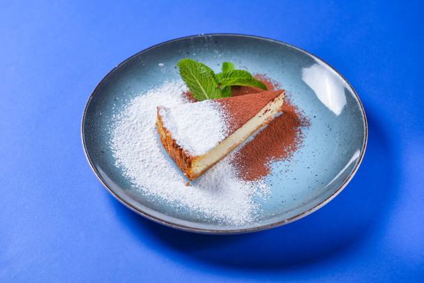 Slice of Plain New York Cheesecake on blue plate over blue background. Piece of cheesecake with sugar and cocoa powder and a leaf of meat. - Φωτογραφία, εικόνα