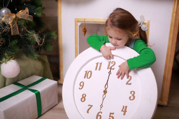 Little girl holding clock near christmas tree. - Foto, imagen