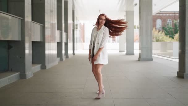 A beautiful happy woman with long red hair in a trendy white coat walks and smiles, rejoicing in her success and freedom. Playful lady goes dancing in downtown. Slow motion.  - Footage, Video