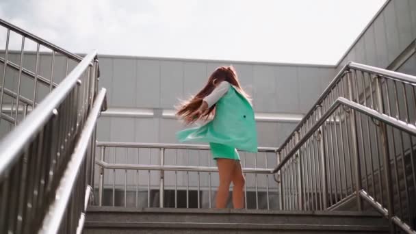 A beautiful slim woman in a trendy turquoise suit is spinning with happiness against the background of the city. Pretty, playful lady rejoices at her success in the downtown area. Slow motion. - Footage, Video