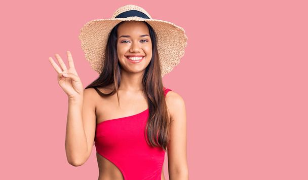 Молодая красивая латиноамериканка в купальниках и летней шляпе показывает и показывает пальцами номер три, улыбаясь уверенно и счастливо.  - Фото, изображение