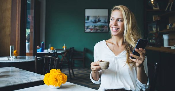 Cheerful woman in white blouse enjoying cup of hot morning coffee while looking away sitting at table and interacting with smartphone in modern coffee house - Foto, immagini