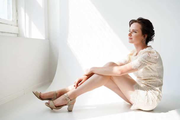 Primo piano di bella giovane donna in abito corto bianco sittinf sul pavimento e guardando nella finestra di luce - Foto, immagini