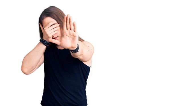 Young adult man with long hair wearing goth style with black clothes covering eyes with hands and doing stop gesture with sad and fear expression. embarrassed and negative concept.  - 写真・画像