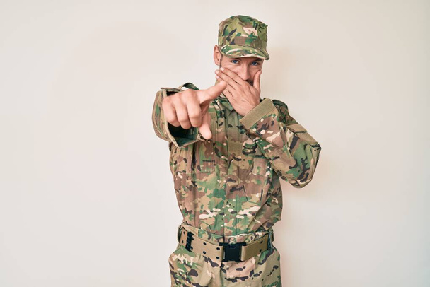 Jonge blanke man in camouflage legeruniform lacht je uit, wijst met de vinger naar de camera met hand over mond, schaamte uitdrukking  - Foto, afbeelding
