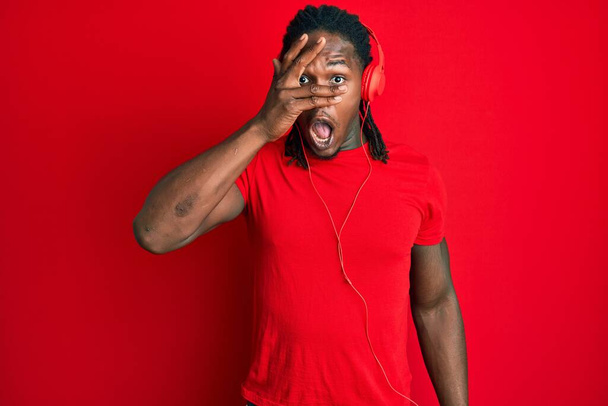 Afro-americano com tranças ouvindo música usando fones de ouvido espreitando em choque cobrindo rosto e olhos com a mão, olhando através dos dedos com medo  - Foto, Imagem
