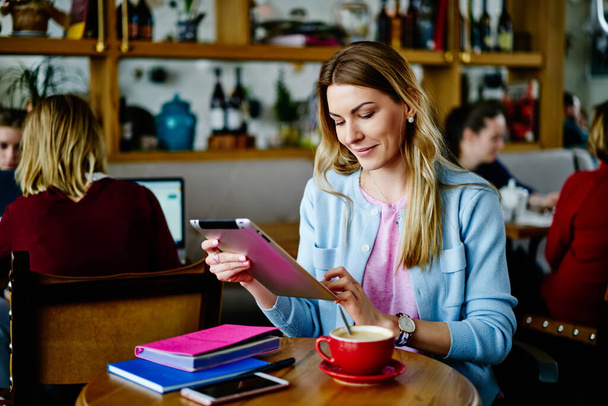 Positive female in casual clothes smiling and browsing browsing tablet while sitting at wooden table with cup of coffee and working in cafe - Φωτογραφία, εικόνα
