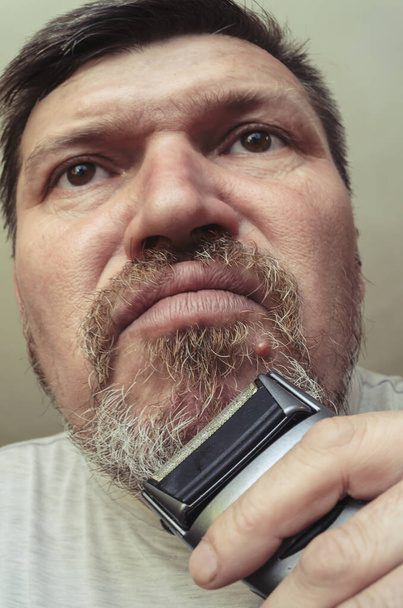 Un homme adulte se rase avec un rasoir électrique. Portrait inhabituel d'un homme barbu aux cheveux gris. Grimaces devant la caméra - Photo, image