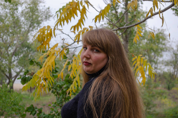 Προσωπογραφία μεσήλικας γυναίκας με ξανθά διαλυμένα μαλλιά σε ένα φθινοπωρινό πάρκο. Γυναίκα σε μαύρο πλεκτό μπλούζα με μακριά ρέοντα μαλλιά. - Φωτογραφία, εικόνα
