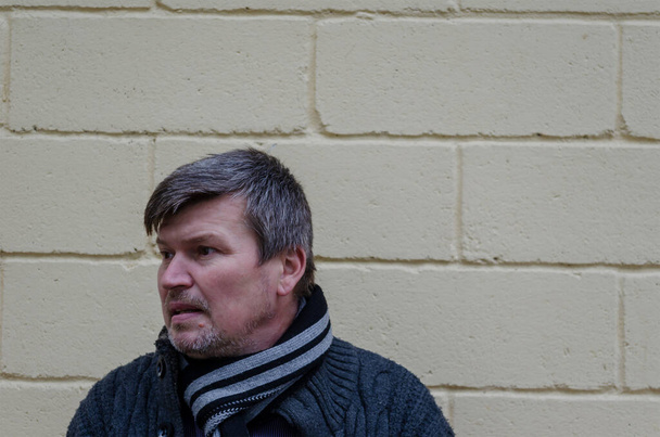 Portrait of man with gray hair looking away at the yellow wall. Middle-aged man in gray sweatshirt and scarf says something to an invisible interlocutor. - Photo, Image