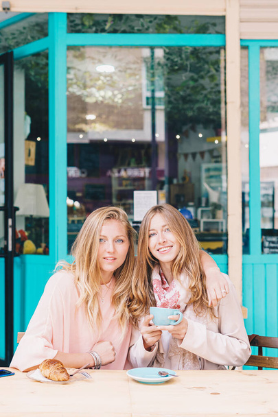 Δύο όμορφα κορίτσια κοιτάζουν την κάμερα ενώ πίνουν καφέ σε μια βεράντα.. - Φωτογραφία, εικόνα