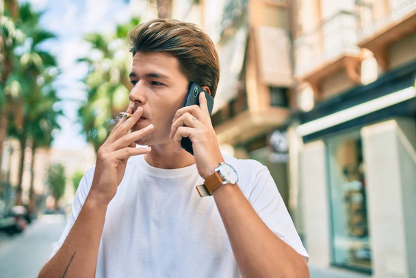 Joven hombre caucásico hablando en el teléfono inteligente y fumar cigarrillos en la ciudad. - Foto, imagen