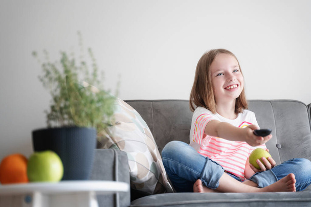 Adorable child girl 8 years old sits on a gray sofa with a green apple and a TV remote control in her hands - 写真・画像