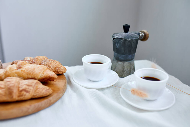 Two coffee cups and Italian coffee maker with croissant over table at home morning breakfast rituals concept, lifestyle food background - Photo, Image