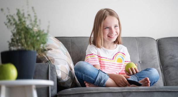 Adorable child girl 8 years old sits on a gray sofa with a green apple and a TV remote control in her hands - Фото, изображение
