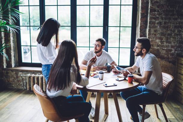 Positieve multiraciale collega 's in casual kleding zitten aan ronde tafel en luisteren naar collega terwijl het verzamelen van zaken in modern kantoor - Foto, afbeelding