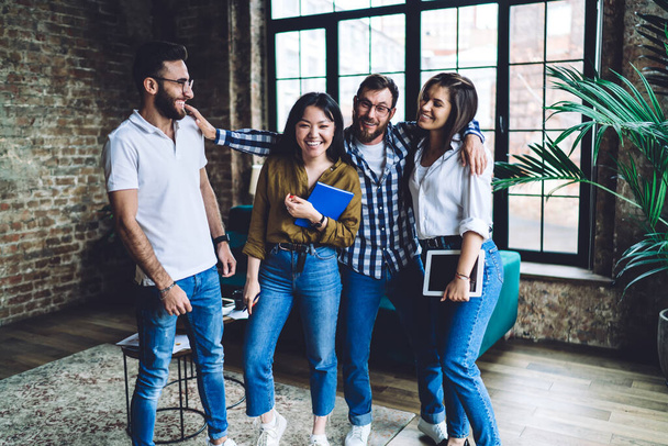 Group of excited multiracial colleagues in casual clothes laughing and hugging while standing together in loft office and chilling during break - Photo, image
