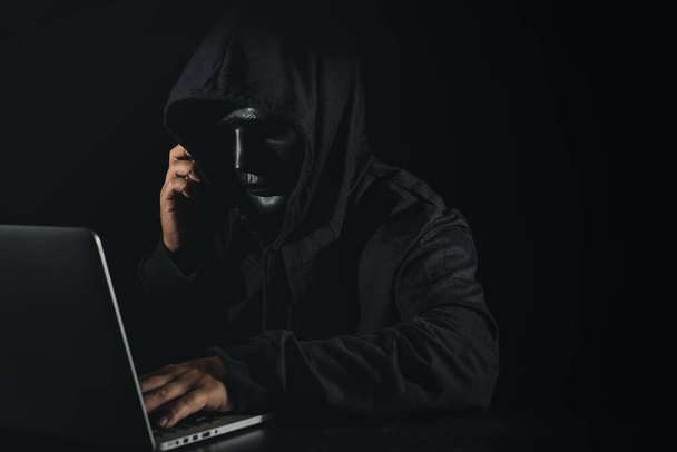 Опасный анонимный хакер в капюшоне и маске, используя компьютер и смартфон на черном фоне, взламывая корпоративный сервер данных безопасности. Интернет-преступление, концепция безопасности кибератак - Фото, изображение