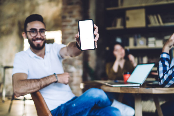 Cheerful guy in glasses showing screen of phone to camera while sitting at table with coworkers in cozy cafe and smiling at camera - Photo, image