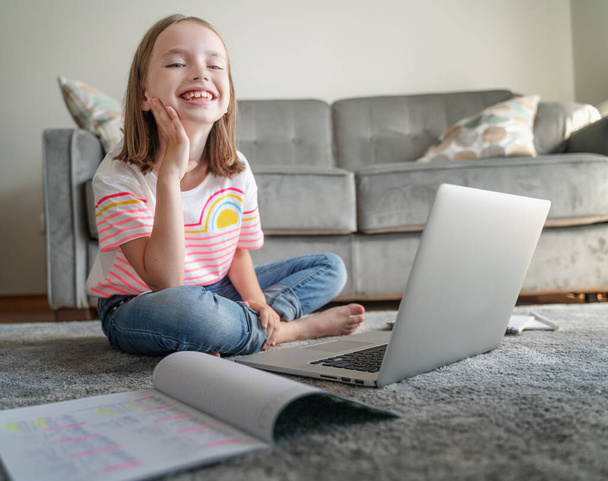 happy smiling beautiful child girl 8 years old sitting on a gray carpet in front of a laptop, homework and online education - Foto, Imagem